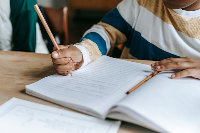 Child writing in a lined notebook with a pencil at a wooden desk, wearing a striped sweater