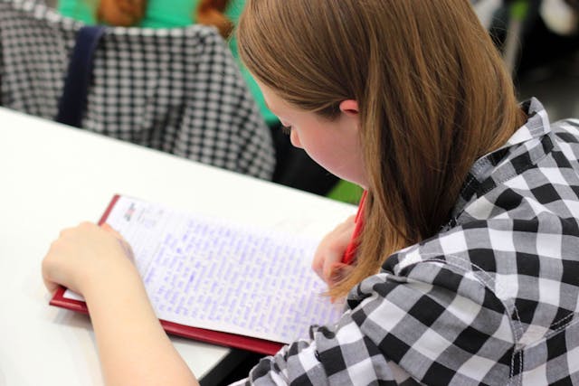 Student writing in a notebook with a red pen at a classroom desk, working on an academic assignment